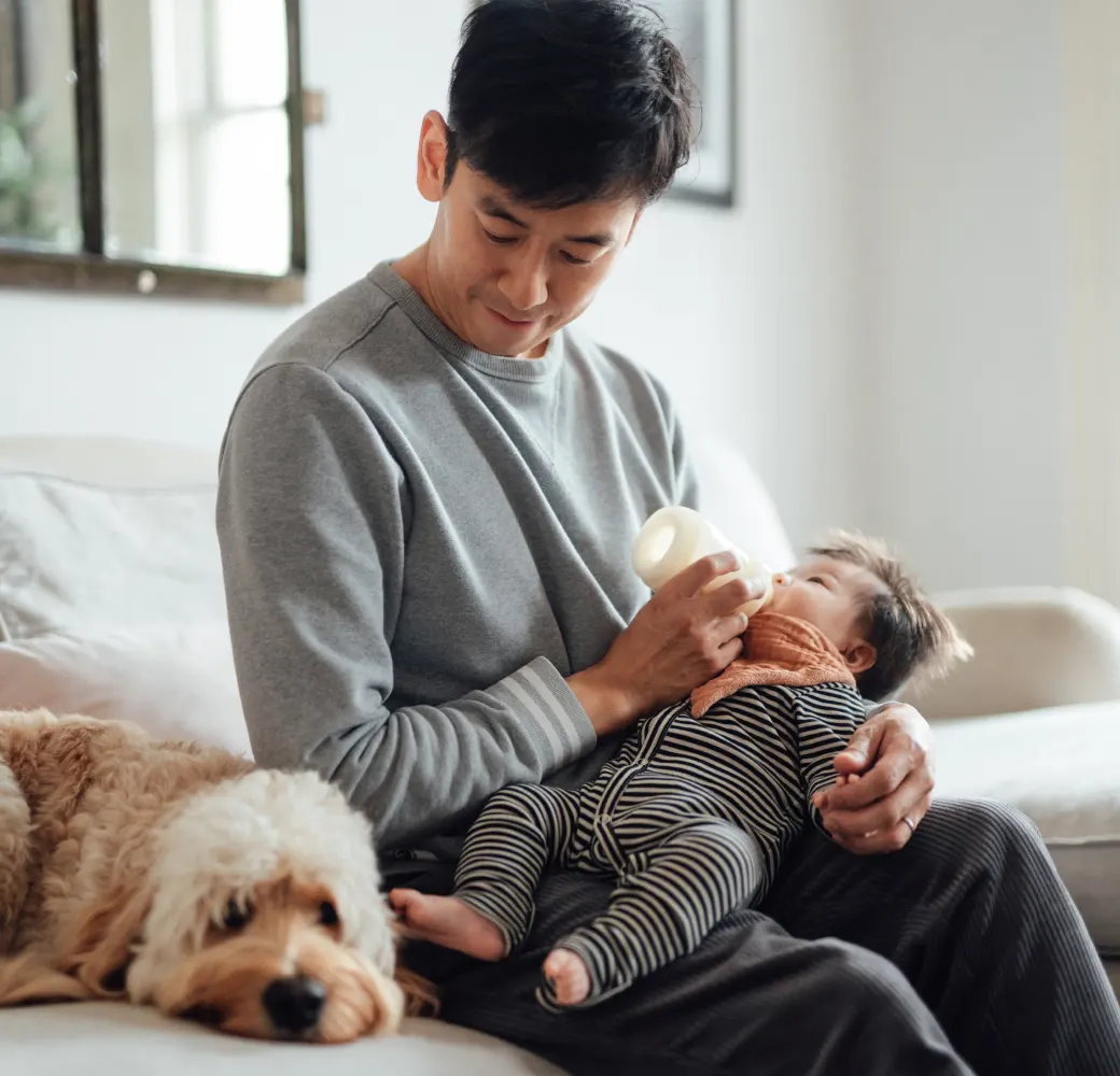Image of a man feeding a baby while sitting next to a dog on a couch