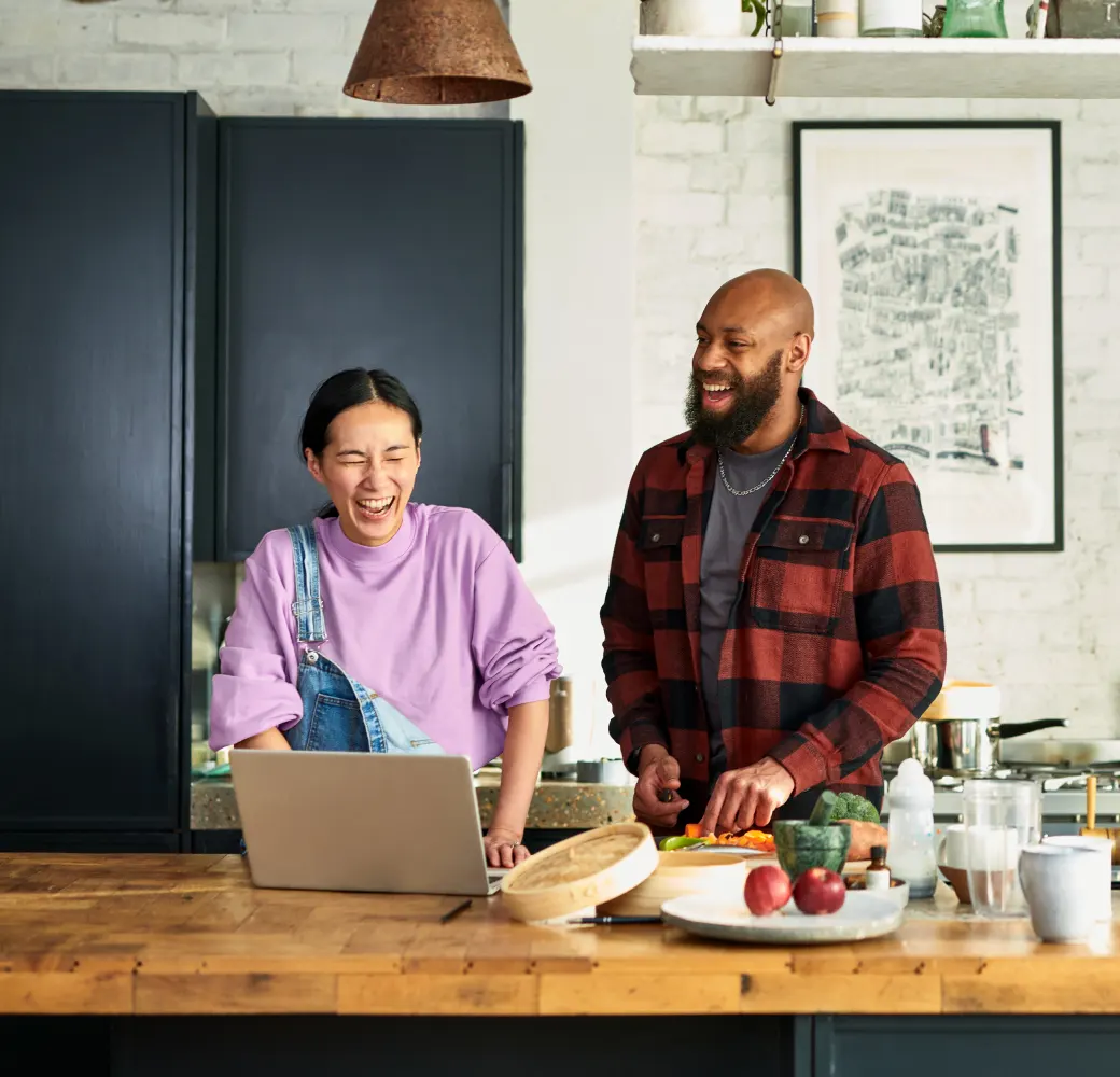 A man and a woman in a kitchen laughing while looking at laptop and preparing food