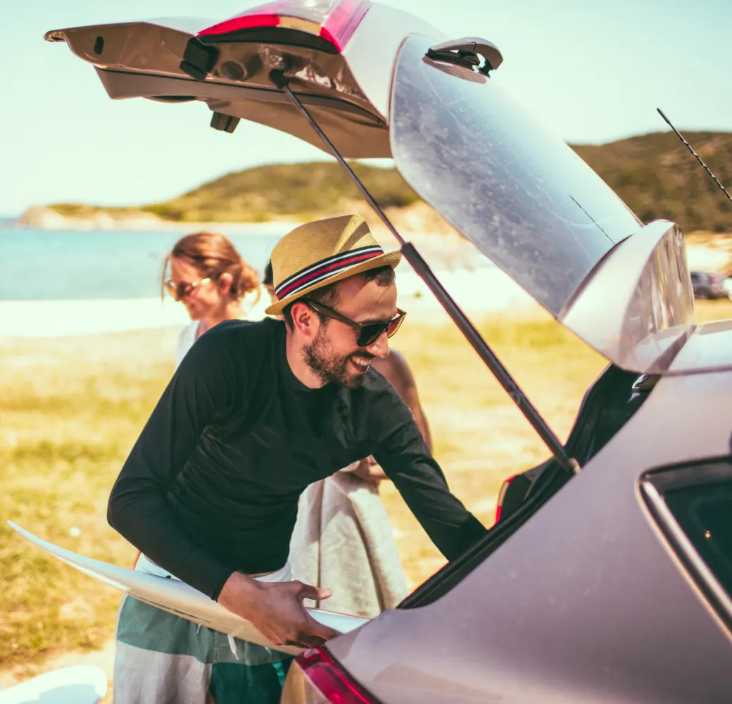 Man pulling a surfboard out of car trunk with woman in the background