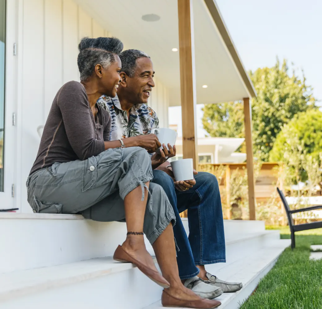 Older couple sitting on the steps of their house drinking coffee looking toward their yard