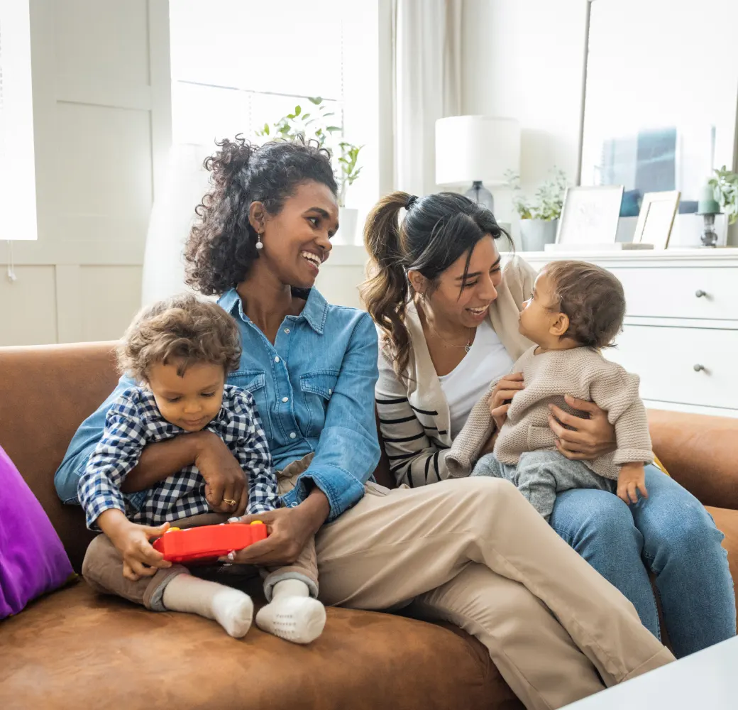 Two women sitting on a couch holding two young children and smiling