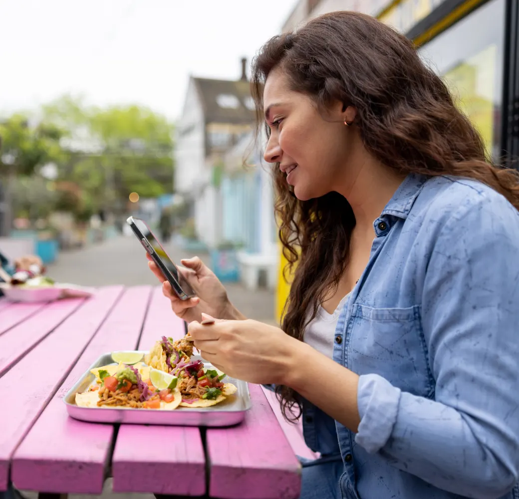 Woman sitting outside at a pink picnic table eating tacos and looking at her phone