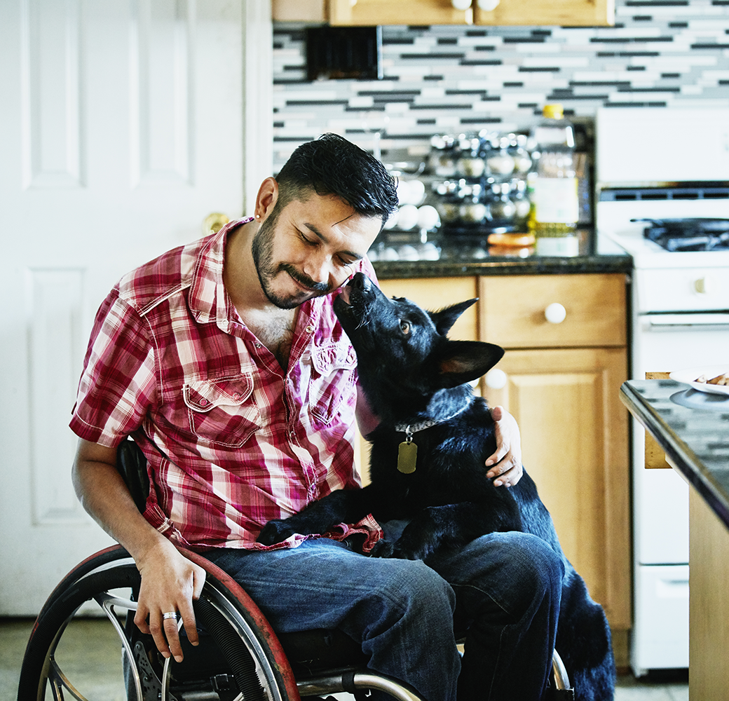Man in wheelchair with black dog kissing him on the cheek
