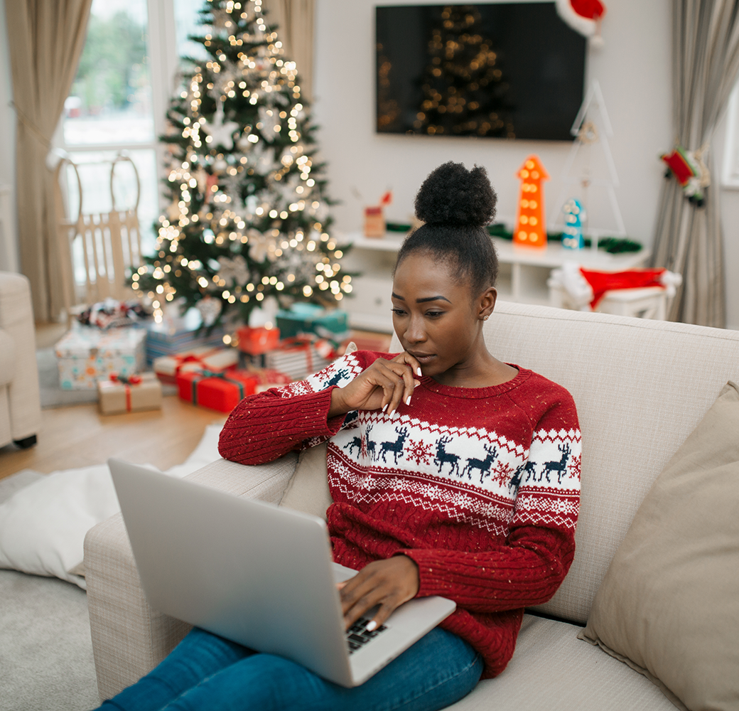 Black woman wearing Christmas sweater sitting on the couch using computer