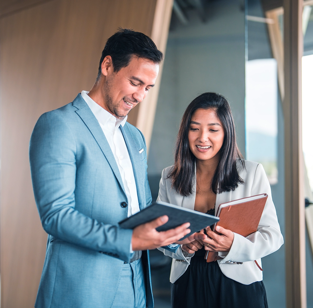 A man and a woman in business attire looking at notebooks and smiling